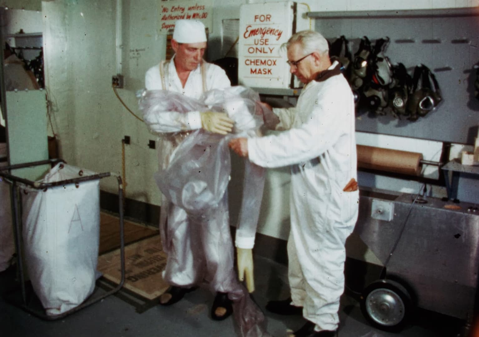 Two men in white suits and gloves holding a plastic bag on a nuclear plant, one assisting the other with a protective suit.